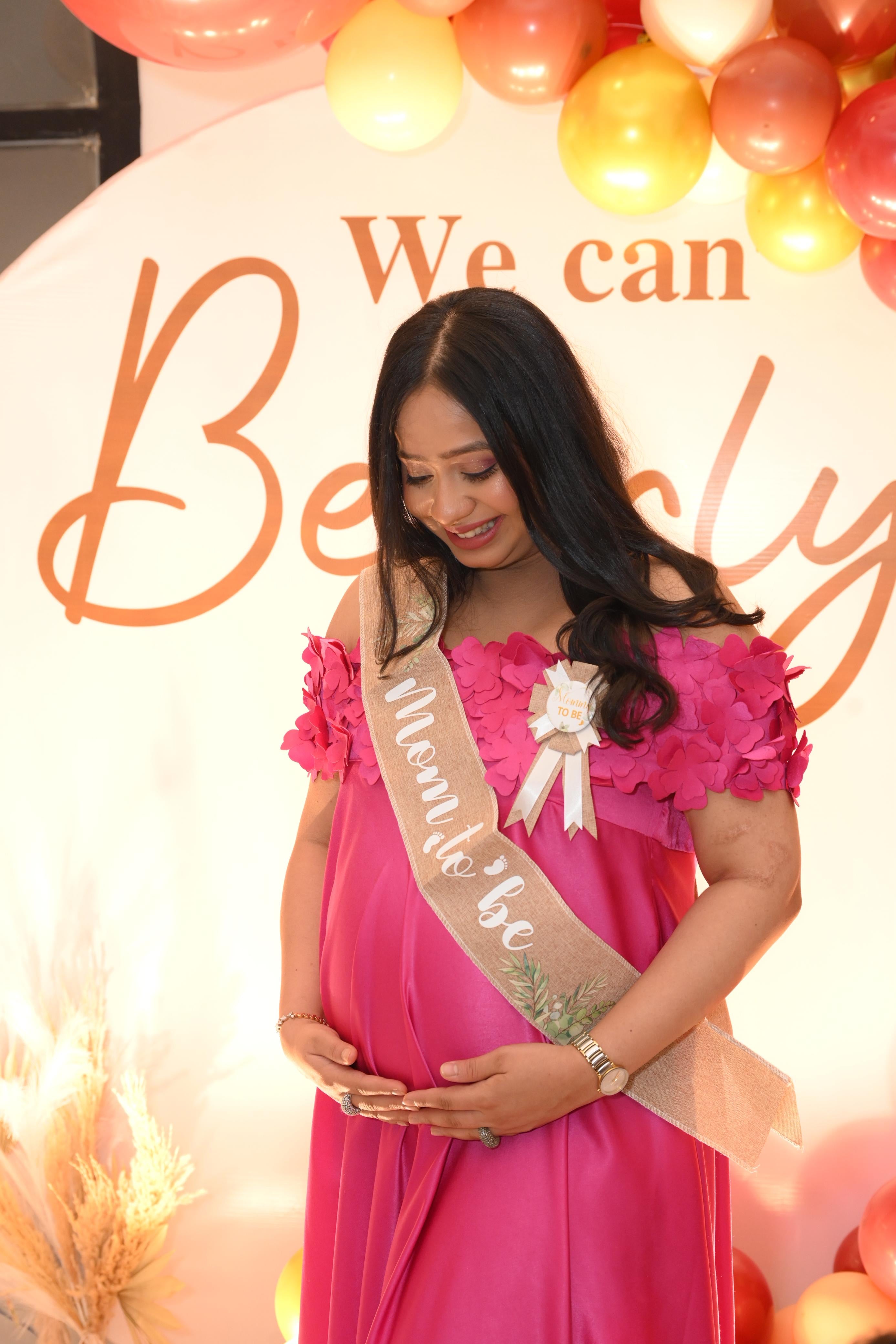 Pregnant woman in a pink dress with a sash standing in front of a decorative backdrop with balloons.